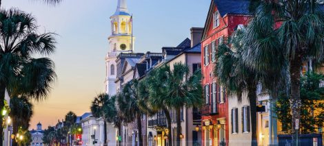 Street view of a historic area at sunset, featuring a row of colorful buildings and palm trees, with a tall church steeple prominently visible in the background.