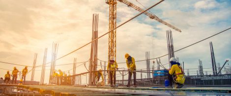 Construction workers wearing safety gear working on a building site with cranes and scaffolding in the background, during sunset.