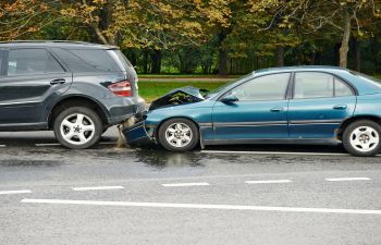 A blue sedan with a damaged front has collided with the rear end of a black SUV on a wet road with trees in the background.