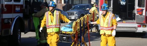 Two firefighters in yellow uniforms and blue helmets carry an empty stretcher on a road near emergency vehicles. Other emergency personnel are in the background.