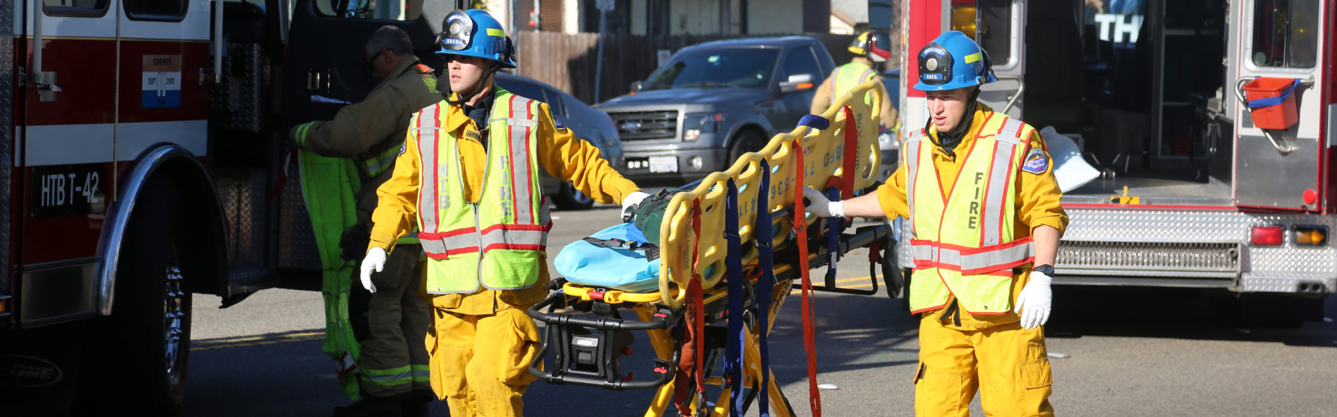 Two firefighters in yellow uniforms and blue helmets carry an empty stretcher on a road near emergency vehicles. Other emergency personnel are in the background.