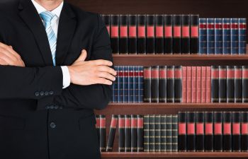 Man in a suit with arms crossed stands in front of a bookcase filled with legal books.