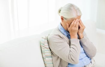 An elderly person with white hair is sitting on a couch, covering their face with both hands. They are wearing a light blue shirt and beige cardigan.