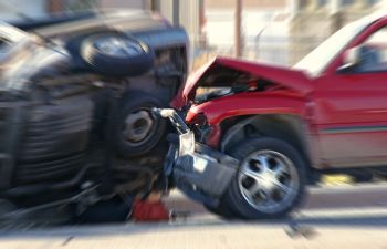 A red SUV collides with a black vehicle that is partially overturned on a street. Both cars display significant damage.