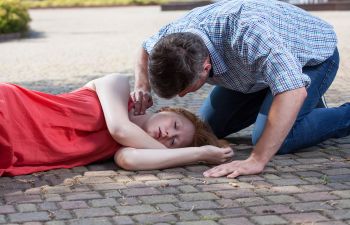 A man kneels beside a woman in a red dress lying unconscious on a paved surface, checking her condition.