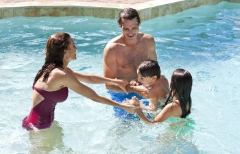 Two adults and two children are standing and playing in a swimming pool, smiling and holding hands.