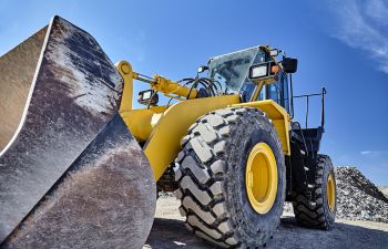A large yellow bulldozer is parked on a construction site, with its shovel raised. The sky is clear and blue in the background.