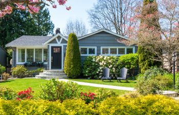 A small grey house with a dark front door, well-maintained lawn, and two chairs in the front garden, surrounded by various trees, bushes, and flowering plants.
