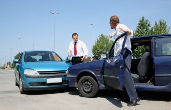 Two men stand beside a blue and a teal car that collided on a clear day. One man checks a clipboard while the other exits his vehicle.