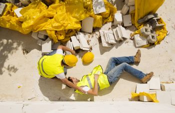 A construction worker is assisting another worker who is lying on the ground amid scattered debris and yellow material, both wearing yellow safety vests and helmets.