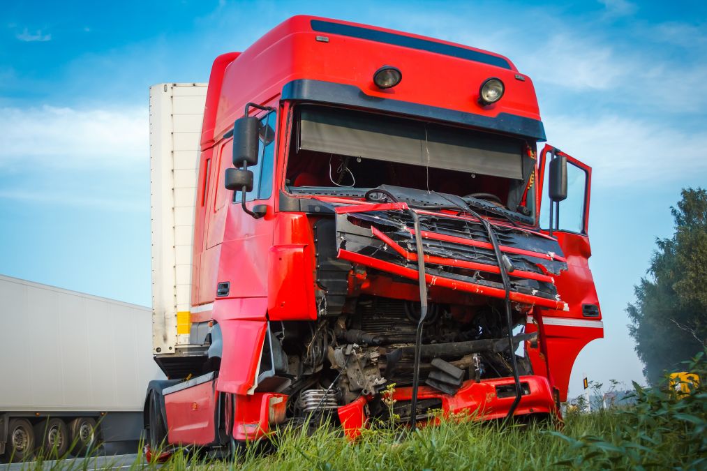 Red truck with a severely damaged front parked on a grassy area. The hood is crumpled and dislodged, exposing the engine. Open driver's door and blue sky in the background.