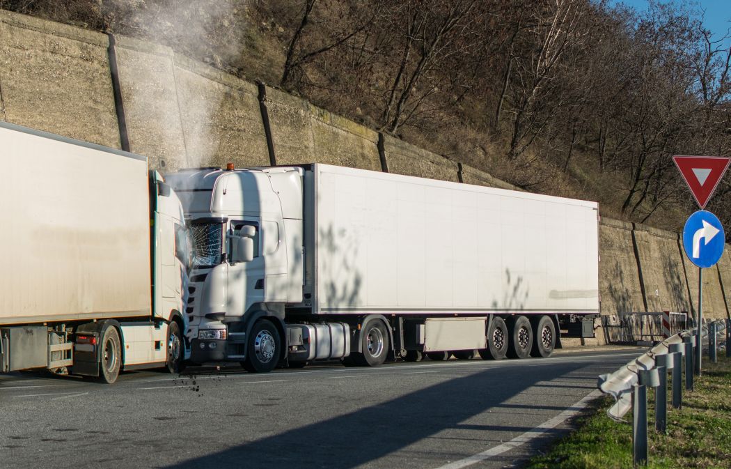Two large white trucks have collided on a roadside with retaining walls. Steam or smoke is visible above the left truck's cab. Road signs are present on the right side.