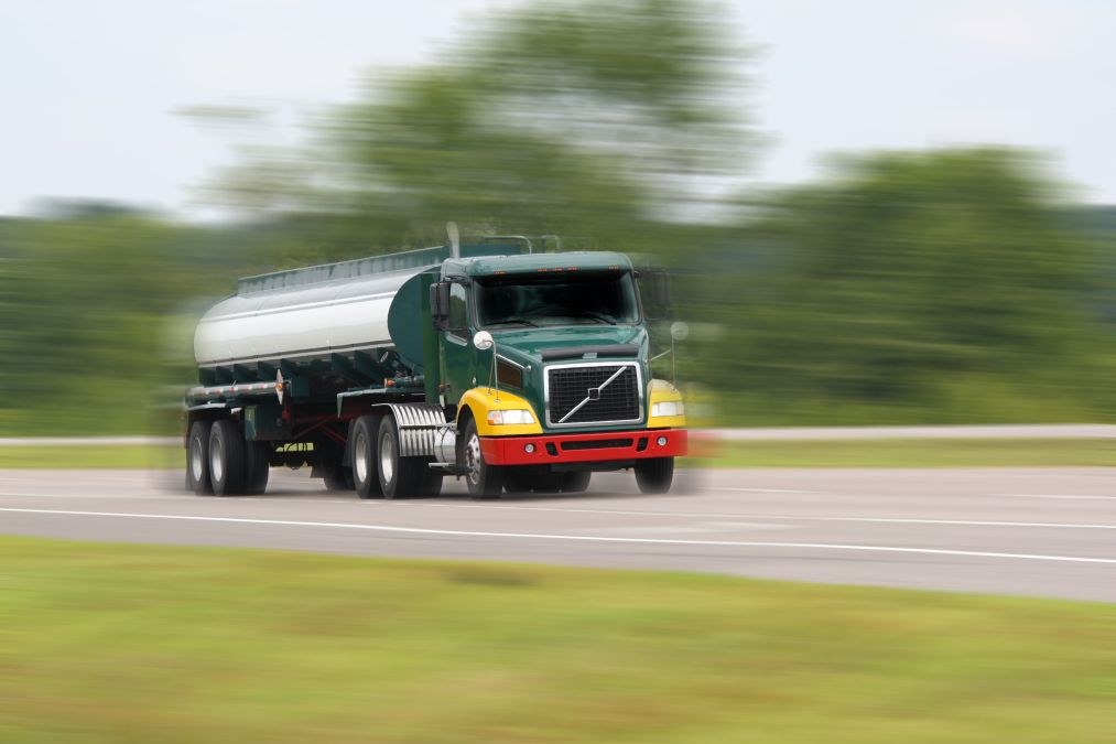 A green and yellow tanker truck speeds down a road, with trees blurred in the background.