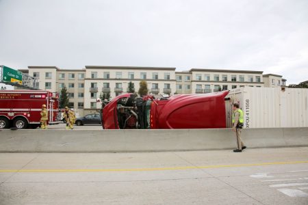 A red truck is overturned on a highway beside a residential building. Firefighters and a fire rescue truck are present at the scene.