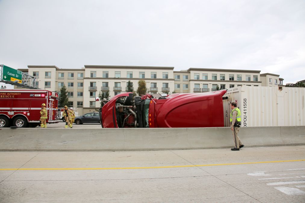 A red truck is overturned on a highway beside a residential building. Firefighters and a fire rescue truck are present at the scene.