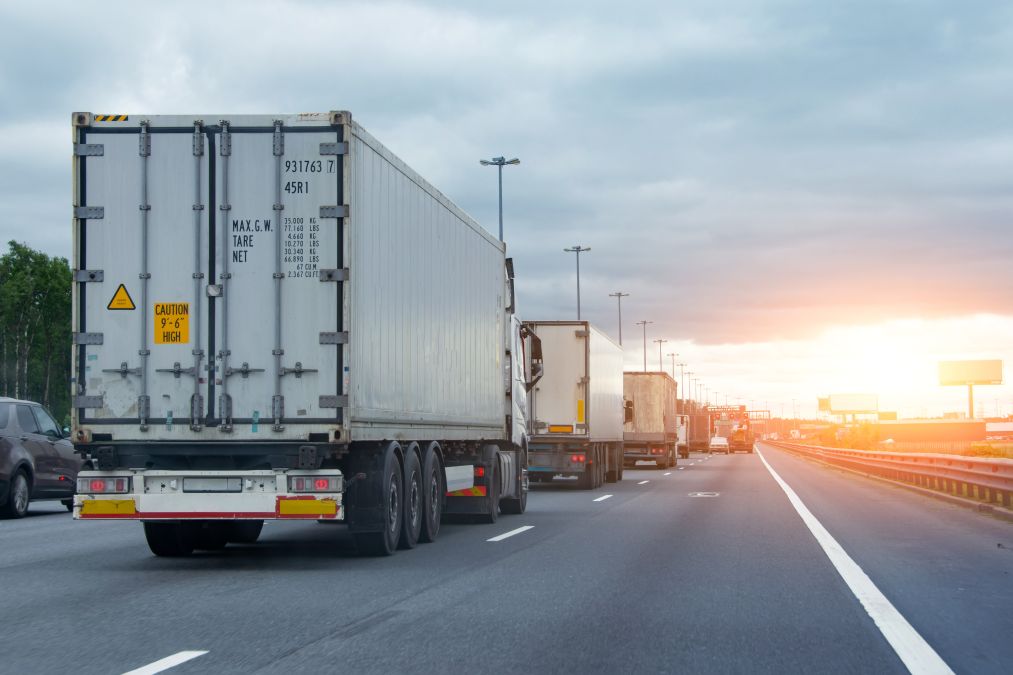 Trucks lined up on a highway, driving towards the sunset. Sky is cloudy, with the road stretching into the distance.