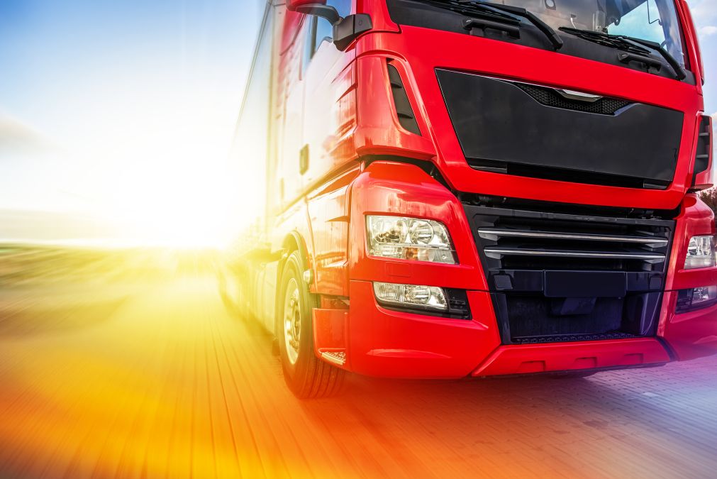 Red truck driving on a road with a bright sun in the background.