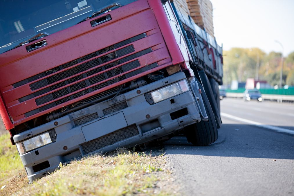 A red truck tilted off the road with its wheels on the grass, carrying a load of timber.   .