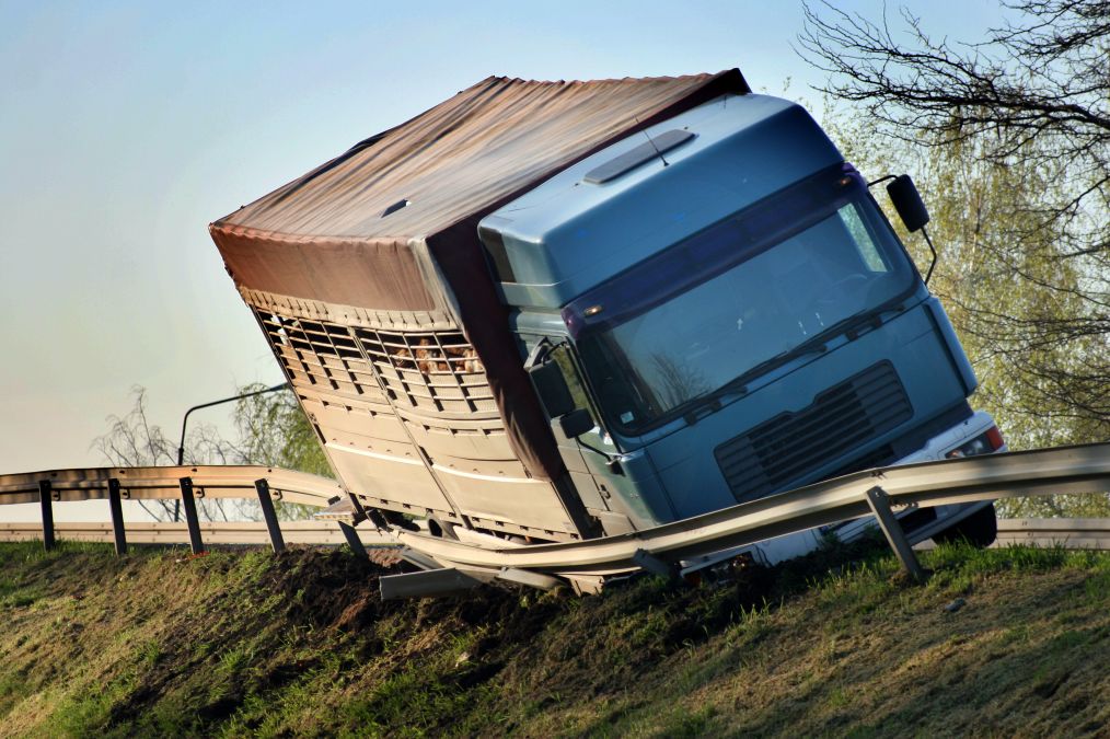 A large truck with a covered cargo area tilts precariously on a grassy roadside, its front end pressed against a bent guardrail.