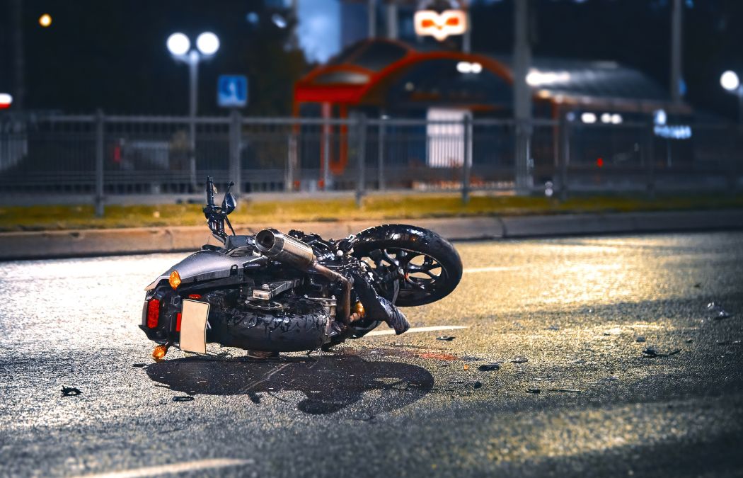 A motorcycle lies overturned on a wet road at night, with debris scattered around it, near an illuminated street and a blurred background of buildings.