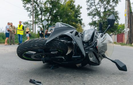 A black motorcycle lies on its side on a road. In the background, people stand near trees and road signs.