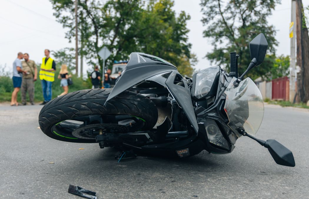 A black motorcycle lies on its side on a road. In the background, people stand near trees and road signs.