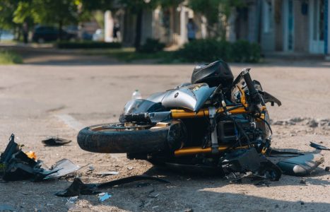 A damaged motorcycle lies on its side on a street, surrounded by debris, under clear daylight conditions.