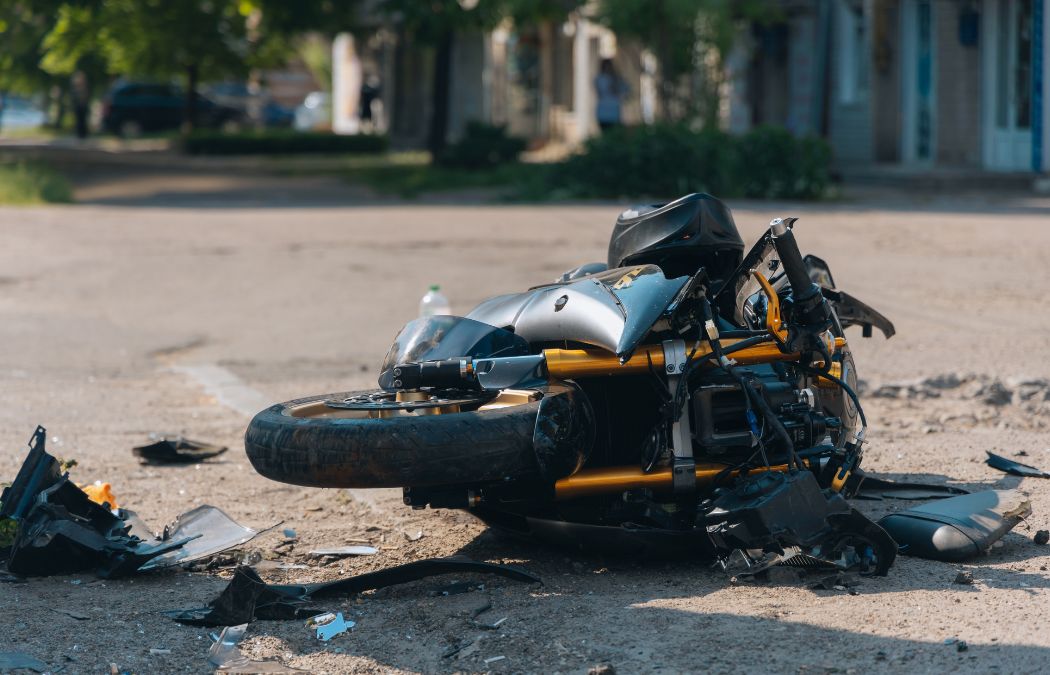 A damaged motorcycle lies on its side on a street, surrounded by debris, under clear daylight conditions.