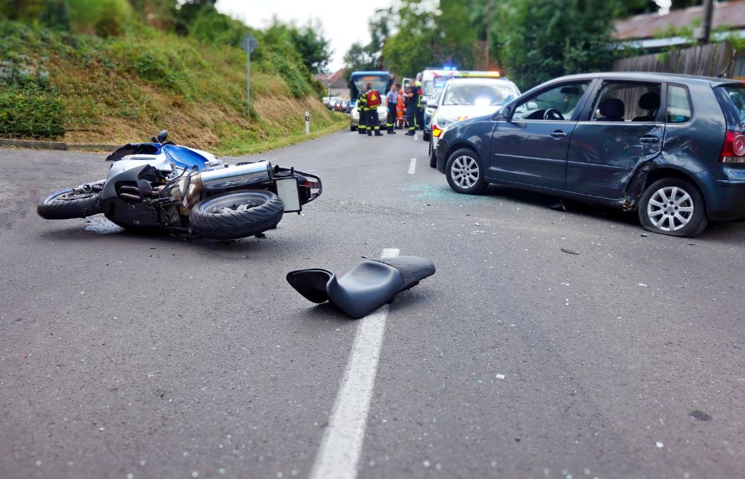 A motorcycle lies on its side near a damaged car on a road. Emergency vehicles and responders are in the background.