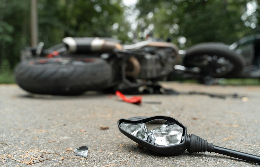 A fallen motorcycle lies on its side on the road with debris scattered, including a broken rearview mirror in the foreground.