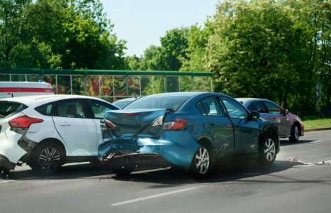 Three cars involved in a rear-end collision on a sunny day, with visible damage to the back and front of the vehicles. Trees and greenery line the background.