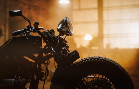 Close-up of a vintage-style motorcycle, with focus on the front wheel and headlight, set against a warmly lit, industrial indoor background with large windows and blurred lights.