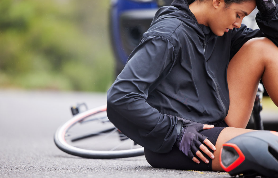 A cyclist sits on the ground next to a fallen bicycle, holding their leg and appearing to be in pain, with a car visible in the background.