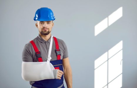 Builder gets injured at work. Portrait of man in workwear uniform, hard hat and sling immobilizer on broken arm standing on copy space wall background. Accident and injury at construction site concept
