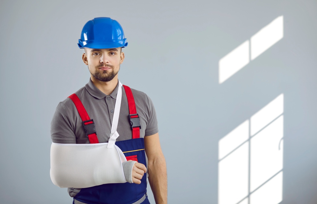 Builder gets injured at work. Portrait of man in workwear uniform, hard hat and sling immobilizer on broken arm standing on copy space wall background. Accident and injury at construction site concept